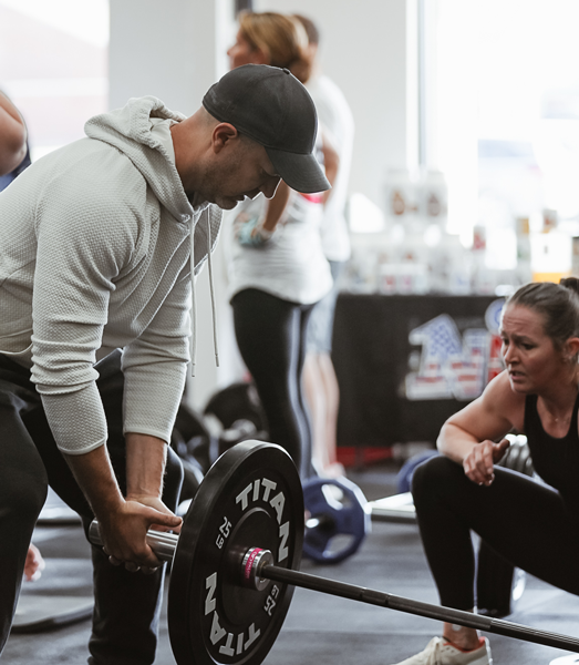 Coach demonstrating proper barbell lifting technique while assisting a member during a group strength training session at Freedom Fitness O’Fallon.