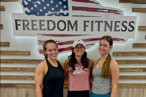Three members posing together in front of the Freedom Fitness O’Fallon logo after a workout session.