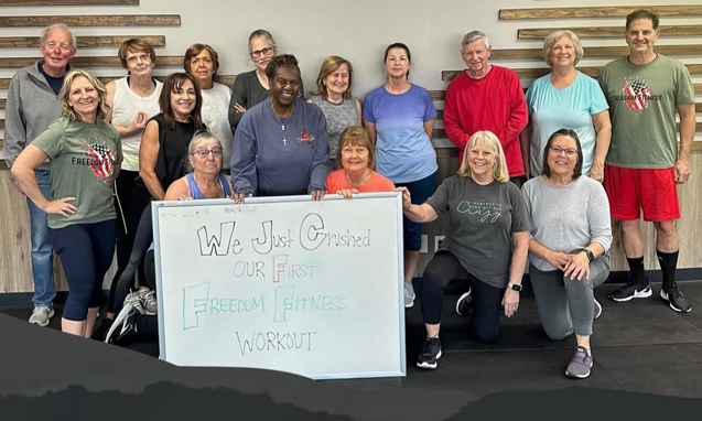 A group of 13 adults posing together inside a fitness studio after completing a workout. Several participants stand behind a whiteboard sign that reads, ‘We Just Crushed Our First Freedom Fitness Workout.’ The group is smiling and dressed in casual athletic clothing, standing on black gym flooring against a wall with wood accent decor.